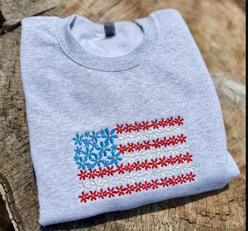 以下が含まれることがあります： 刺繍されたアメリカ国旗のデザインが施された、明るい灰色のスウェットシャツ。旗の星は青い花で、ストライプは赤と白の花でできています。スウェットシャツは柔らかい素材で作られています。