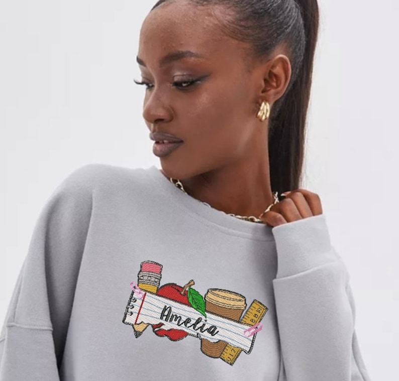 May include: Gray sweatshirt with a graphic of a red apple, pencil, ruler, and coffee cup. The graphic also includes the text "Amelia" on a white banner.