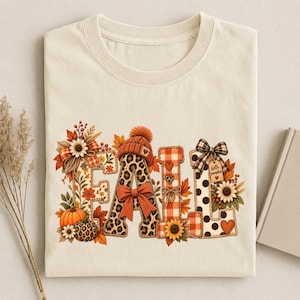 Op de afbeelding: Een crèmekleurig t-shirt met het woord FALL in grote letters. De letters zijn versierd met luipaardprint, oranje ruitjes en zwarte stippen, geaccentueerd met illustraties van pompoenen, zonnebloemen, herfstbladeren en een muts.