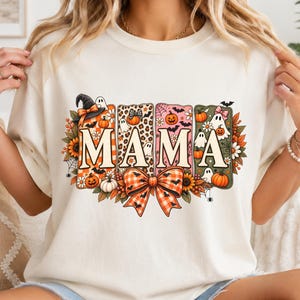 May include: A cream-coloured t-shirt featuring a graphic print with the word MAMA in large letters. Each letter is framed by Halloween-themed designs including ghosts, pumpkins, bats, spiderwebs, sunflowers, and a large orange plaid bow.