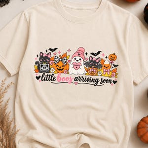 Puede incluir: Una camiseta de color crema con un gráfico colorido de Halloween que muestra un gato negro con una ecografía, una calabaza, un fantasma con gorro rosa, un murciélago y un dulce de maíz. El texto dice little boos arriving soon.