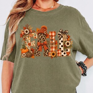May include: An olive green short-sleeve t-shirt featuring the word FALL in large block letters. The letters are decorated with patterns including leopard print, orange plaid, and polka dots, accented with pumpkins, sunflowers, and a beanie.
