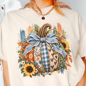 Könnte beinhalten: Ein cremefarbenes T-Shirt mit einer Grafik eines Kürbisses, der mit einer großen blauen Schleife, Sonnenblumen und verschiedenen Mustern wie blauem Vichy-Karo, Blumenprints und Herzen verziert ist. Das Design zeigt herbstliche Blätter und Beeren.