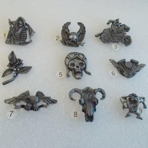 May include: Nine silver-toned metal pins with various designs, including a skull with wings, a skull with a motorcycle helmet, a rose, a skull with horns, and a cartoon character. The pins are numbered 1 through 9.