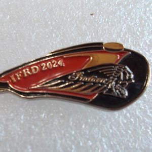 May include: A metal lapel pin shaped like a motorcycle fuel tank, featuring a black, red, and gold color scheme. The pin displays the text IFRD 2024 in gold lettering on a red background, alongside a gold Indian Motorcycle logo graphic.