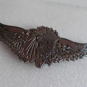 Könnte beinhalten: Detailliertes Metallabzeichen mit dem Kopf eines indianischen Häuptlings mit Federkopfschmuck, flankiert von stilisierten Flügeln. Die Worte "Indian Motorcycles" sind in Rot auf die Flügel geschrieben. Das Abzeichen hat ein metallisches Finish.