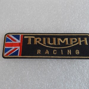 K&ouml;nnte beinhalten: Schwarzer rechteckiger Aufn&auml;her mit goldfarbenem gesticktem Text "TRIUMPH RACING" und einer Union Jack-Flagge auf der linken Seite. F&uuml;r Motorradfans.