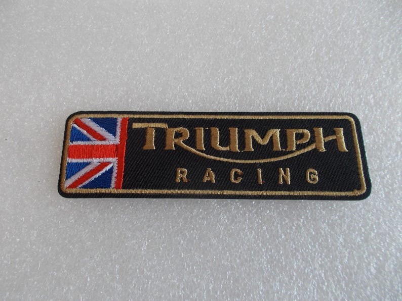 K&ouml;nnte beinhalten: Schwarzer rechteckiger Aufn&auml;her mit goldfarbenem gesticktem Text "TRIUMPH RACING" und einer Union Jack-Flagge auf der linken Seite. F&uuml;r Motorradfans.