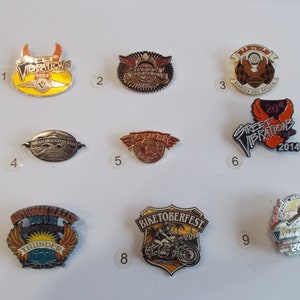 Può includere: Una collezione di nove spille a tema motociclistico. Le spille presentano vari disegni, tra cui ali, motociclette e testo come "Street Vibrations 2014" e "Biketoberfest 2014". Le spille sono in vari colori, tra cui oro, argento, rosso e nero.
