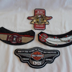 May include: Four embroidered patches featuring Harley Davidson logos. The top patch is red with a white eagle and the text "15th Anniversary Harley Owners Group 1983-1998". The second patch is black with a red and white Harley Davidson logo and the text "2001". The third patch is black with a white feather and the text "HOG 2005". The bottom patch is black with a silver Harley Davidson logo and the text "Ninety Five Years of Great Motorcycles 1903 95 1998".
