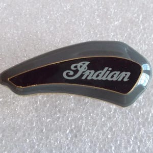 May include: A curved, teardrop-shaped emblem featuring a glossy gray outer border and a black central inlay. The word Indian is printed in a white, stylized script font across the black section, accented by a thin gold-colored outline.