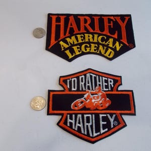 May include: Two Harley-Davidson patches. The top patch reads "HARLEY AMERICAN LEGEND" in orange and yellow on a black background. The bottom patch features a motorcycle and the words "I'D RATHER HARLEY" in orange and white on a black background.