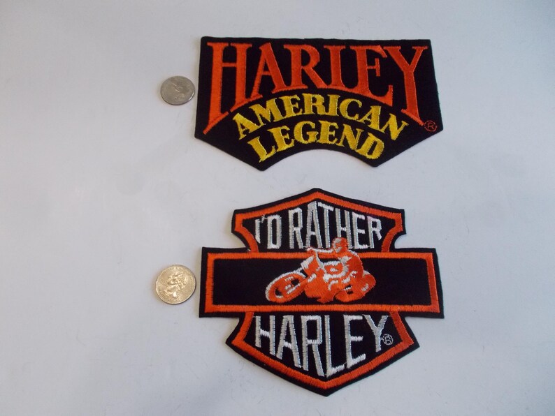 May include: Two Harley-Davidson patches. The top patch reads "HARLEY AMERICAN LEGEND" in orange and yellow on a black background. The bottom patch features a motorcycle and the words "I'D RATHER HARLEY" in orange and white on a black background.