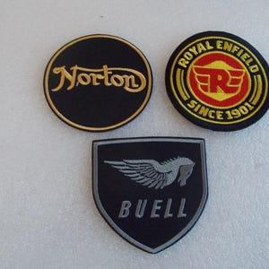 May include: Three embroidered patches featuring motorcycle brand logos. One circular black patch with gold Norton text, one circular black patch with red and yellow Royal Enfield text, and one black shield-shaped patch with a grey winged horse and Buell text.