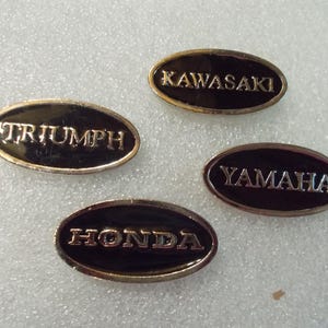 May include: Four oval-shaped metal emblems with a black enamel finish and gold-toned borders. Each emblem features a raised metallic brand name in capital letters: Triumph, Kawasaki, Yamaha, and Honda. The emblems are arranged in a scattered layout.
