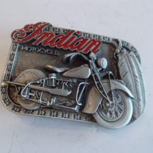 Puede incluir: Una hebilla de cinturón de plata con una imagen detallada de una Indian Motorcycle. La hebilla incluye las palabras "Indian Motorcycle" y una imagen estilizada de una motocicleta con plumas.