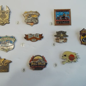 Op de afbeelding: Een verzameling van tien motorfiets-evenementenpins. De pins zijn meestal goud en zilver met kleurrijke ontwerpen. Sommige van de pins bevatten de woorden "Biketoberfest", "Bike Week" en "H.O.G."
