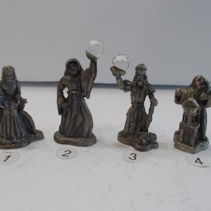 May include: Four pewter figurines of wizards, each holding a crystal ball. The wizards are wearing robes and have long beards. The figurines are on a white surface.