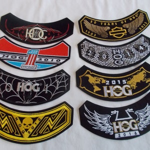 May include: A collection of black, curved embroidered patches. Designs include Harley-Davidson logos, years, and the text "HOG". Patches feature various colors, including red, gold, and white, with intricate details and lettering.