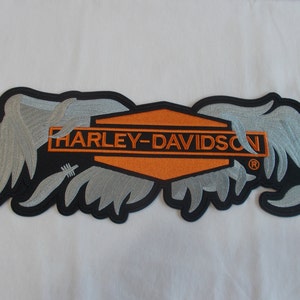 May include: A black and orange embroidered patch with the Harley-Davidson logo and wings. The wings are silver and the logo is surrounded by a black border.