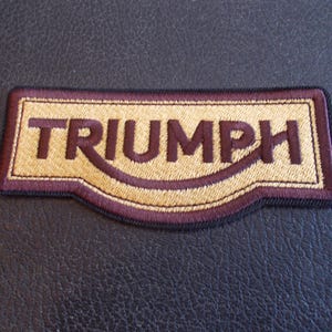 May include: Embroidered patch featuring the word "TRIUMPH" in dark brown lettering on a tan background. The patch has a burgundy border and a black outline. The patch is rectangular with a curved bottom edge.