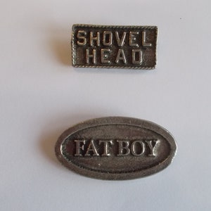 Puede incluir: Dos pines de metal plateados con texto grabado. El primer pin es rectangular y dice "SHOVEL HEAD". El segundo pin es ovalado y dice "FAT BOY".