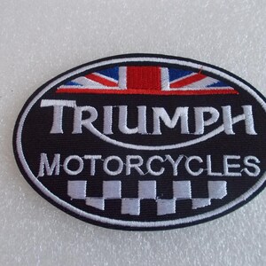 K&ouml;nnte beinhalten: Ovaler Triumph Motorcycles-Aufn&auml;her mit schwarzem Hintergrund, wei&szlig;em Rand und wei&szlig;em Text. Die Oberseite zeigt ein Union Jack-Design in Rot, Wei&szlig; und Blau. Der Boden hat ein Schachbrettmuster.