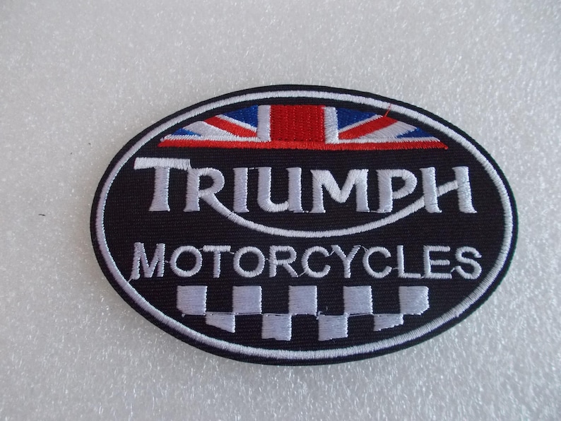 K&ouml;nnte beinhalten: Ovaler Triumph Motorcycles-Aufn&auml;her mit schwarzem Hintergrund, wei&szlig;em Rand und wei&szlig;em Text. Die Oberseite zeigt ein Union Jack-Design in Rot, Wei&szlig; und Blau. Der Boden hat ein Schachbrettmuster.