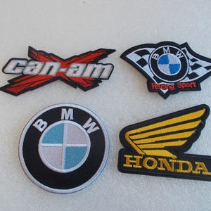 May include: Four embroidered patches featuring automotive logos. Top left is a red and silver Can-Am logo. Top right is a BMW Racing Sport logo with checkered flags. Bottom left is a circular BMW logo. Bottom right is a yellow Honda wing logo.