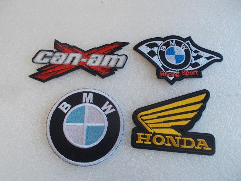 Pode incluir: Quatro emblemas bordados com logotipos automotivos. No canto superior esquerdo, um logotipo Can-Am vermelho e prateado. No canto superior direito, um logotipo BMW Racing Sport com bandeiras quadriculadas. Abaixo, um logotipo circular da BMW e um logotipo de asa da Honda amarelo.