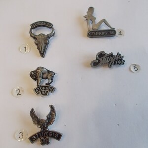 Könnte beinhalten: Fünf silberne Sturgis Motorcycle Rally Pins. Die Pins zeigen einen Büffel, einen Adler, einen Schädel, eine Frau und das Sturgis-Logo. Die Pins sind von 1 bis 5 nummeriert.