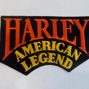 May include: Black patch with orange embroidered text reading "HARLEY" above "AMERICAN LEGEND" in yellow. The patch has a unique shape with a curved bottom edge.
