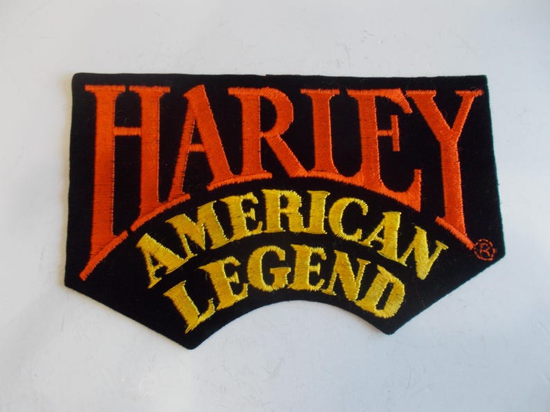 May include: Black patch with orange embroidered text reading "HARLEY" above "AMERICAN LEGEND" in yellow. The patch has a unique shape with a curved bottom edge.