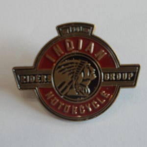 May include: A circular metal pin featuring a red and black enamel design with the text INDIAN MOTORCYCLE RIDERS GROUP. The center displays a profile illustration of a person wearing a feathered headdress, rendered in a metallic bronze tone.
