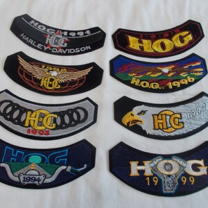 May include: Collection of Harley-Davidson HOG patches in various colors. Each patch features the HOG logo and year, with designs including eagles, motorcycles, and the Harley-Davidson name. Patches are black, blue, and silver.