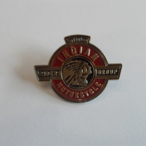 May include: A circular pin with a red and gold design. The pin features the words "Indian Motorcycle Riders Group" and an image of a Native American chief. The pin is likely a collectible item or emblem.