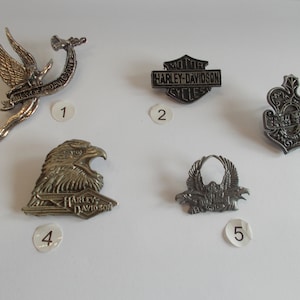 May include: Five silver and black Harley Davidson motorcycle pins. The pins feature different designs, including an eagle, a winged angel, and the Harley Davidson logo.