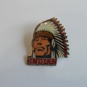 May include: A gold-colored pin with a colorful image of a Native American man wearing a headdress. The text "INDIAN" is printed below the image.