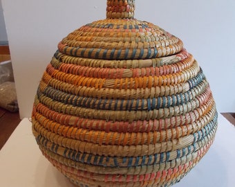 May include: A round, woven basket with a lid. The basket is made of natural materials and has a colorful striped pattern in shades of orange, blue, and green.