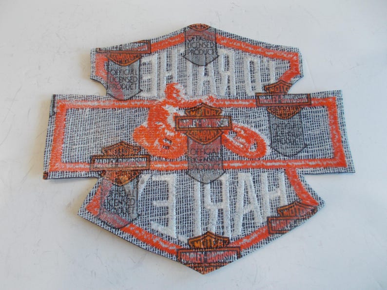 May include: A Harley-Davidson patch with a textured, gray background and orange and white accents. The patch features the Harley-Davidson logo and the words "HARLEY-DAVIDSON" and "OFFICIAL LICENSED PRODUCT".