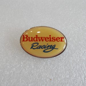 May include: Oval Budweiser Racing badge with a yellow background and a red and blue logo. The text "Budweiser" is in red, and "Racing" is in blue, both in a stylized font. The badge has a metallic border.