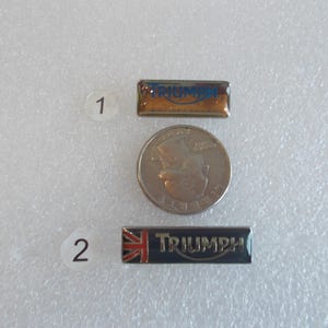 May include: Two metal pins with the word "Triumph" in blue lettering on a gold and black background. The pins are numbered 1 and 2.