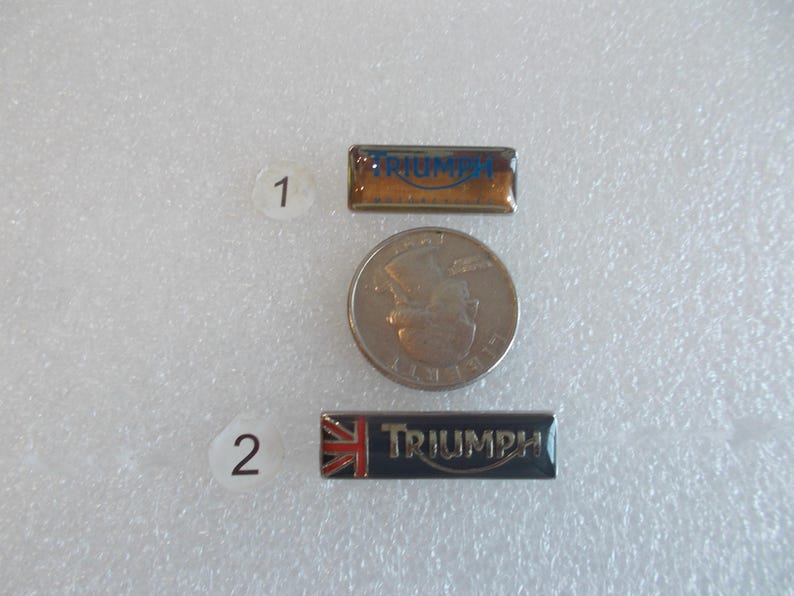 May include: Two metal pins with the word "Triumph" in blue lettering on a gold and black background. The pins are numbered 1 and 2.
