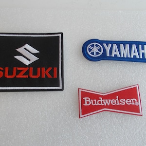 May include: Three embroidered patches featuring brand logos. The Suzuki patch is black with a white logo and red text. The Yamaha patch is blue with a white logo and text. The Budweiser patch is red with white text.