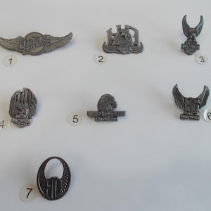 Może przedstawiać: Siedem srebrnych pinów Harley Davidson z różnymi wzorami, w tym skrzydlatym logo, orłem i napisem Harley Davidson.