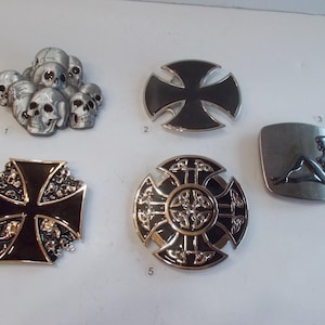 May include: Collection of metal belt buckles with various designs. Includes a cluster of skulls, a black cross, a square buckle with a reclining woman, and two cross-shaped buckles with ornate detailing. The buckles are silver and black.