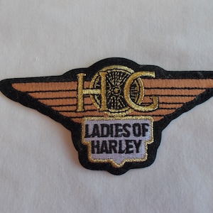 May include: A black and gold embroidered patch with a winged Harley Davidson logo and the text "Ladies of Harley".