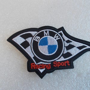 Pode incluir: Um emblema bordado com o log&oacute;tipo da BMW em azul, branco e preto, ladeado por bandeiras de corrida axadrezadas a preto e branco. O texto Racing Sport est&aacute; bordado com fio vermelho na parte inferior do emblema, que tem um rebordo preto.