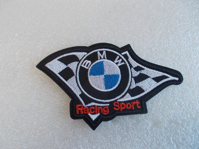 Pode incluir: Um emblema bordado com o log&oacute;tipo da BMW em azul, branco e preto, ladeado por bandeiras de corrida axadrezadas a preto e branco. O texto Racing Sport est&aacute; bordado com fio vermelho na parte inferior do emblema, que tem um rebordo preto.