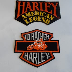 May include: Two Harley-Davidson patches. The top patch reads "HARLEY AMERICAN LEGEND" in orange and yellow lettering on a black background. The bottom patch features a motorcycle graphic and the word "HARLEY".